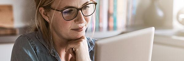 Seguro de Proteccion de Pagos - Mujer con gafas sentada en la cocina de su casa con una tablet en la mano y papeles sobre la mesa, trabajando online Seguro de Proteccion de Pagos - Mujer con gafas sentada en la cocina de su casa con una tablet en la mano y papeles sobre la mesa, trabajando online